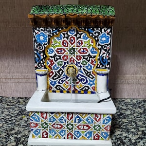 May include: A decorative ceramic fountain with intricate geometric patterns in red, blue, yellow, and green. The fountain has a white basin and a green tiled roof. A golden spout dispenses water.