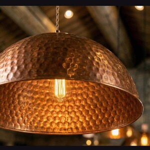 May include: A copper pendant light with a hammered texture. The dome-shaped shade houses a vintage-style Edison bulb, emitting a warm, inviting glow. The light hangs from a twisted cord, perfect for a kitchen or dining room.