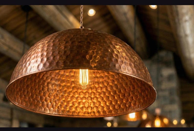 May include: A copper pendant light with a hammered texture. The dome-shaped shade houses a vintage-style Edison bulb, emitting a warm, inviting glow. The light hangs from a twisted cord, perfect for a kitchen or dining room.