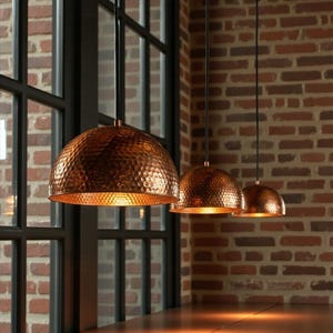 May include: Three copper pendant lights with a hammered texture hang above a wooden surface. The lights are dome-shaped and suspended by black cords. A brick wall and a large window frame the scene, creating a warm, inviting ambiance.