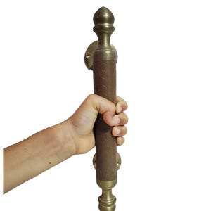 Könnte beinhalten: Ein Türgriff aus Messing mit einem braunen Ledergriff. Der Türgriff hat dekorative Details und wird von einer Hand vor weißem Hintergrund gehalten. Die Messingkomponenten haben eine polierte Oberfläche.