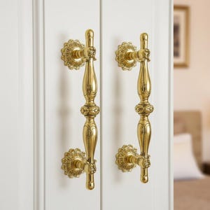 Op de afbeelding: Twee sierlijke, goudkleurige deurgrepen gemonteerd op een witte deur. Elke handgreep heeft een decoratieve bloemenrozet aan de basis en een gedetailleerde, langwerpige greep. De handgrepen zijn symmetrisch.