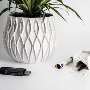 May include: A white ceramic planter with a unique wavy design, featuring a green plant with long leaves. A white key holder with a key inside, a rolled-up piece of paper, and a black USB card reader are also visible on the white surface.