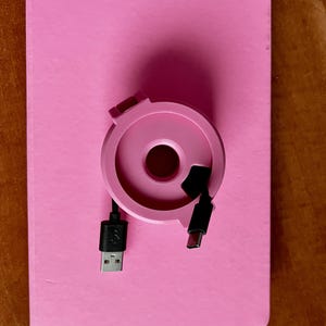 Könnte beinhalten: Ein rosa Kabelorganizer mit einem schwarzen USB-Kabel und einem schwarzen USB-C-Kabel. Der Organizer befindet sich auf einem rosa Notizbuch.