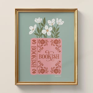 May include: A framed art print featuring a pink book with the word "BOOKISH" in a decorative font. White flowers and green leaves emerge from the book. The background is a muted teal color, and the frame is gold.