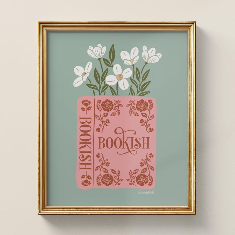 May include: A framed art print featuring a pink book with the word "BOOKISH" in a decorative font. White flowers and green leaves emerge from the book. The background is a muted teal color, and the frame is gold.
