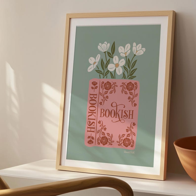 May include: A framed art print featuring a pink book with the word "BOOKISH" and floral accents. White flowers with green leaves sit atop the book against a muted green background. The frame is light wood.