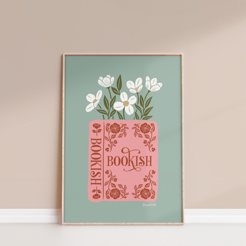 May include: A framed art print featuring a pink book with the word "BOOKISH" in a decorative font. White flowers with green leaves are arranged above the book. The background is a muted teal color.