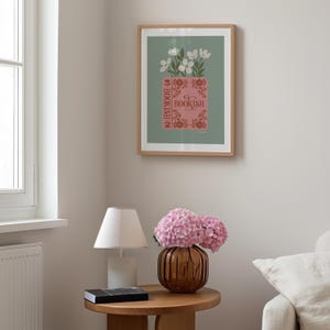 May include: A framed art print featuring a pink book with the word "BOOKISH" and white flowers. The print is in a light wood frame and hangs on a light green background. The room also includes a table lamp, a vase with pink flowers, and a small round table.