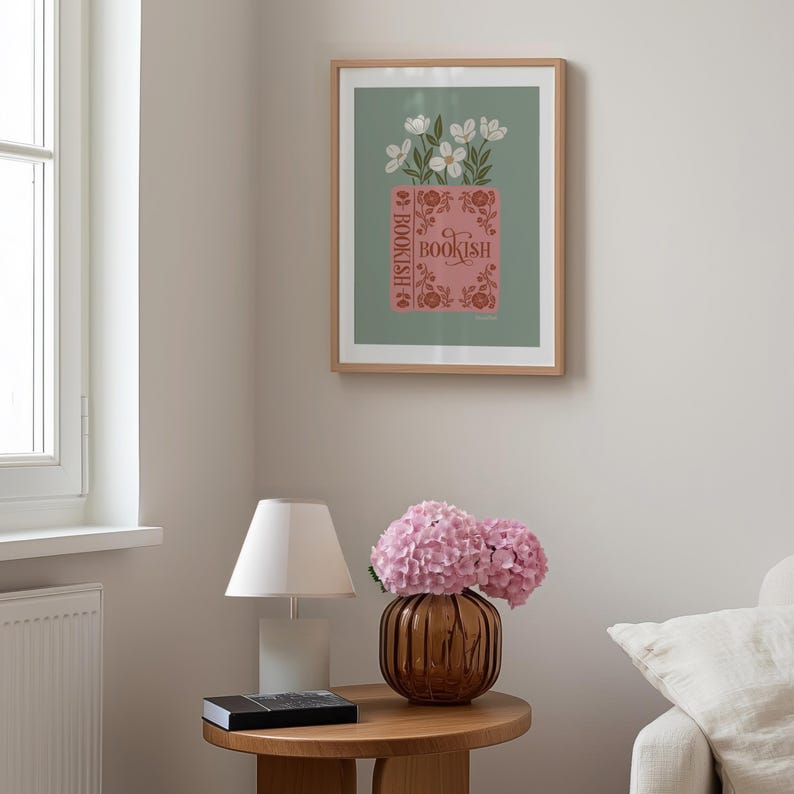 May include: A framed art print featuring a pink book with the word "BOOKISH" and white flowers. The print is in a light wood frame and hangs on a light green background. The room also includes a table lamp, a vase with pink flowers, and a small round table.
