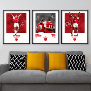 May include: Three framed prints showcasing Arsenal football legends Tony Adams, Dennis Bergkamp, Thierry Henry, and Ian Wright. Each print has a red backdrop with player images, signatures, and names. The prints are displayed above a grey sofa with yellow and black cushions.