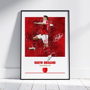 May include: A digital art print of a soccer player in a red and white uniform, with a red background and white paint splatters. The player is in mid-air, kicking the ball. The player's name, "MARTIN ØDEGAARD", is printed below the image, along with the team name and number, "Team Name | #8".