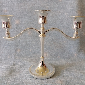 Può includere: Un candelabro d'argento a tre bracci. Ogni portacandela ha la forma di una coppa e poggia su bracci curvi che si estendono da un fusto centrale. La base è rotonda con un bordo decorativo. Il candelabro è realizzato in metallo lucido e riflettente.