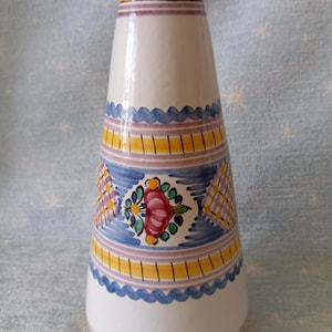May include: A conical ceramic vase with a white base, featuring a yellow rim and base. The surface is decorated with horizontal bands of blue wavy lines, yellow stripes with vertical marks, and purple lines, centered with a floral motif.