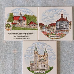 Könnte beinhalten: Drei quadratische Keramikfliesen mit detaillierten Illustrationen deutscher Stadtlandschaften. Eine Fliese zeigt ein Gebäude mit rotem Dach und Text, eine andere einen Stadtplatz und die letzte eine Kirche mit zwei Türmen. Die Fliesen sind vor einem hellblauen Hintergrund angeordnet.