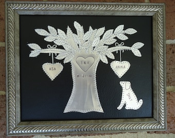 Puede incluir: Un árbol de metal plateado con tres corazones colgando de las ramas. Los corazones están etiquetados como "Asa", "Anika" y "E & K". El tronco del árbol tiene la fecha "8-21-10" inscrita en él. Una silueta de perro de metal plateado está debajo del árbol, etiquetada como "Shay". La obra de arte está enmarcada en un marco plateado.