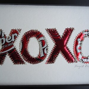 K&ouml;nnte beinhalten: Gerahmtes Kunstwerk mit den Buchstaben "xoxo" aus recycelten Dr. Pepper-Dosen. Die roten und silbernen Buchstaben sind mit silbernem Faden auf einem wei&szlig;en Hintergrund zusammengen&auml;ht. Die Signatur des K&uuml;nstlers ist sichtbar.