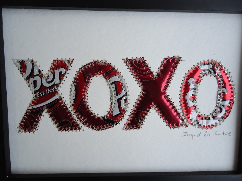 K&ouml;nnte beinhalten: Gerahmtes Kunstwerk mit den Buchstaben "xoxo" aus recycelten Dr. Pepper-Dosen. Die roten und silbernen Buchstaben sind mit silbernem Faden auf einem wei&szlig;en Hintergrund zusammengen&auml;ht. Die Signatur des K&uuml;nstlers ist sichtbar.