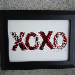 K&ouml;nnte beinhalten: Gerahmtes Kunstwerk mit den Buchstaben "XOXO", hergestellt aus recycelten Dr. Pepper-Dosen. Die rot-wei&szlig;en Buchstaben sind auf einem wei&szlig;en Hintergrund aufgen&auml;ht und in einem schwarzen Rahmen eingefasst. Das Kunstwerk enth&auml;lt den Text "Ingrid Mc. Intire".