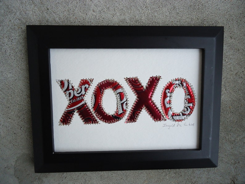K&ouml;nnte beinhalten: Gerahmtes Kunstwerk mit den Buchstaben "XOXO", hergestellt aus recycelten Dr. Pepper-Dosen. Die rot-wei&szlig;en Buchstaben sind auf einem wei&szlig;en Hintergrund aufgen&auml;ht und in einem schwarzen Rahmen eingefasst. Das Kunstwerk enth&auml;lt den Text "Ingrid Mc. Intire".