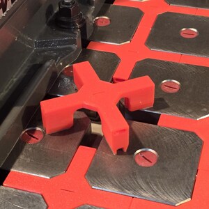 May include: A bright red, X-shaped plastic spacer tool positioned on a grid of square metal plates. The tool features four legs designed to fit between the plates, which are set into a red base with circular red inserts in each corner.