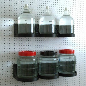 May include: Three glass jars with red lids and three glass jugs with black lids are mounted on a white pegboard wall using black plastic brackets. The jars and jugs are filled with a dark liquid.