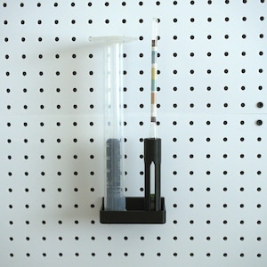 Può includere: Un supporto in plastica nera con un cilindro graduato trasparente e un kit di test del pH in vetro. Il supporto è montato su una bacheca bianca forata.