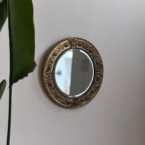 Puede incluir: Un pequeño espejo redondo con un marco dorado y ornamentado. El marco presenta un diseño detallado con patrones repetidos. El espejo está montado en una pared blanca, con una planta verde visible en el fondo.