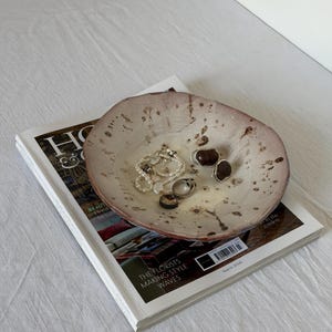 Puede incluir: Un cuenco de cerámica poco profundo, formado a mano, con un borde irregular y ondulado. El cuenco presenta un interior de color crema con motas marrones y un exterior de color marrón rosáceo. Dentro del cuenco hay varias joyas, incluyendo una pulsera.