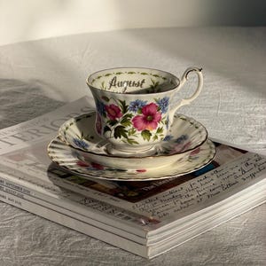 May include: A floral teacup and saucer set with gold trim, resting on a stack of magazines. The teacup is adorned with pink and blue flowers and the word "August". The set is placed on a white tablecloth.