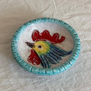 May include: A white ceramic dish with a blue rim and a hand-painted rooster design. The rooster has a red comb, yellow eyes, blue and green feathers, and a white beak.