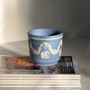 Könnte beinhalten: Hellblaue Keramikschale mit weißen Reliefdetails, darunter eine Girlande und klassische Figuren. Die Schale steht auf einem Stapel Zeitschriften mit dem Titel "The Art of Living Beautifully".