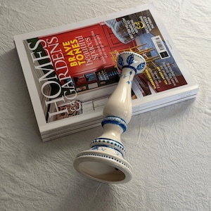 May include: A cream ceramic candlestick with blue floral patterns, placed on a stack of magazines. The magazine cover displays the words "HOMES GARDENS" and "BRAVE TONES beautiful spaces".