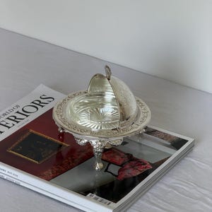 Puede incluir: Una mantequera plateada con tapa abovedada, detalles ornamentados y tres patas decorativas. El plato está abierto, revelando un interior de cristal transparente. La mantequera está sobre una revista con el texto "WORLD OF INTERIORS".