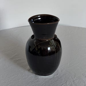 Puede incluir: Un jarrón de cerámica marrón oscuro con un acabado brillante. El jarrón tiene un cuerpo bulboso y una parte superior abierta y ensanchada. Dos pequeñas asas redondeadas están en los lados. El jarrón probablemente se utiliza con fines decorativos o para contener flores.