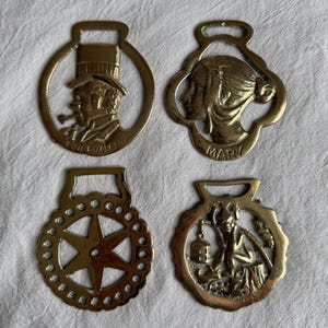 May include: Four antique brass horse brasses. The brasses feature various designs, including a man with a pipe, a woman with the name "Mary", a starburst, and a figure with a lantern. Each brass has a loop at the top for hanging.