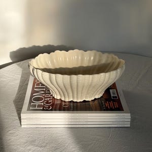 May include: A decorative cream-coloured ceramic bowl, shaped like a shell, with a textured surface. It rests on a stack of "Homes & Gardens" magazines. The magazines display the text "The Art of Living Beautifully" and the months March, July, August, and September.