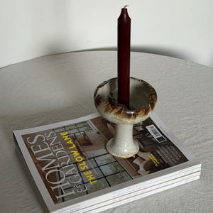 May include: A ceramic candle holder with a dark red candle sits atop a stack of "Homes & Gardens" magazines. The candle holder has a speckled, off-white base and a brown-edged top. The magazines are stacked neatly.