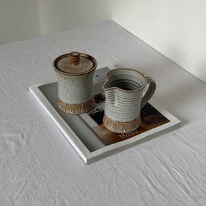 May include: A ceramic sugar bowl with a lid and a matching creamer pitcher. Both pieces feature a speckled light gray glaze with a textured brown band at the base decorated with a repeating square pattern. The creamer has a handle and a spout.