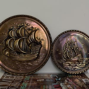 May include: Two circular wall plaques with sailing ship designs. The larger plaque is bronze-coloured, showing a detailed ship. The smaller plaque has a similar ship design with an iridescent finish, both are decorative.