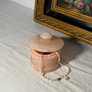 Könnte beinhalten: Eine pfirsichfarbene Schmuckschatulle mit Deckel und kleinem Knopf. Eine weiße Perlenkette mit silbernen Akzenten hängt aus der geöffneten Schatulle. Die Schatulle steht auf einer weißen Oberfläche, mit einem gerahmten Gemälde im Hintergrund.