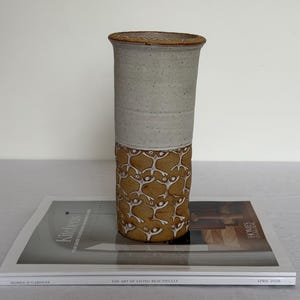 May include: A tall cylindrical ceramic vase with a two-tone finish. The upper portion is a smooth, speckled light gray, while the lower section features a textured, earthy brown surface decorated with a repeating pattern of small white human figures.