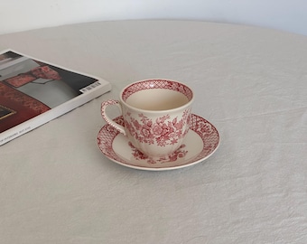 Può includere: Un set di tazza e piattino bianchi con un motivo floreale rosso. La tazza ha un manico curvo e poggia su un piattino abbinato. Il set è posto su una superficie bianca, con una rivista visibile sullo sfondo.