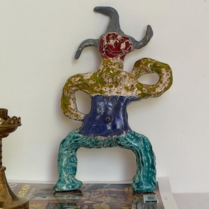 Puede incluir: Una colorida escultura de cerámica de una figura con rostro rojo, cuernos grises y cuerpo verde y azul. La figura está posando con los brazos levantados y las piernas separadas. La escultura se exhibe sobre una pila de revistas.