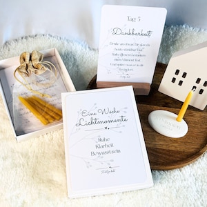 Op de afbeelding: Een witte doos met kaarsen, kaarten met Duitse tekst, een klein wit huisje en een kaarsenhouder. De kaarten bevatten zinnen als "Eine Woche Lichtmomente" en "Tag 5 Dankbarkeit".