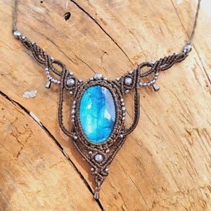 May include: A handcrafted macrame necklace featuring a large, oval-shaped, blue gemstone pendant. The pendant is surrounded by intricate brown macrame knots and silver-colored beads. The necklace is displayed on a wooden surface.