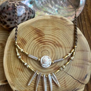 Può includere: Una collana artigianale con un ciondolo a conchiglia a spirale e una frangia di pezzi di conchiglia allungati. La collana ha un cordino marrone e dettagli metallici color oro. Il design è ambientato su uno sfondo di legno.