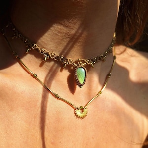 Könnte beinhalten: Eine mehrlagige Halskette mit einem grünen Edelstein-Anhänger und einem goldenen Sonnenstrahlen-Anhänger. Die Halskette besteht aus braunem Band und goldenen Perlen.