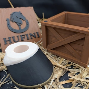 Könnte beinhalten: Eine braune Holzkiste, ein hufeisenförmiges Objekt mit schwarzen und weißen Akzenten und ein braunes Schild mit einem Pferdekopf- und Hufeisenlogo sowie dem Wort "HUFINI". Die Objekte befinden sich auf einem Strohbett.