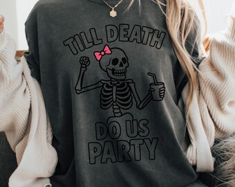 May include: Dark gray t-shirt with the text "Till Death Do Us Party" and a skeleton graphic with a pink bow. The skeleton is holding a drink. The shirt is a casual style.